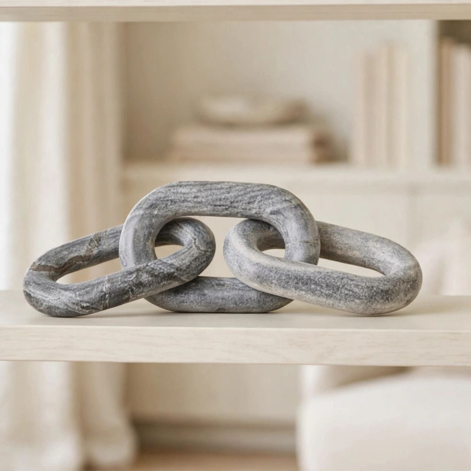 A decorative sculpture made of three interlocking textured gray links, displayed on a light wood shelf in a softly blurred interior setting.