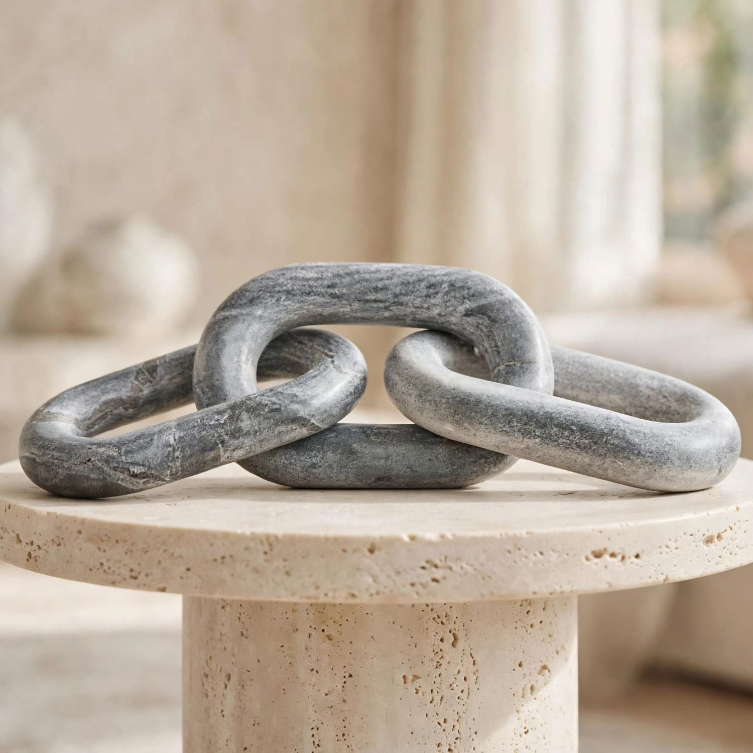 A gray stone sculpture of interlocking links resting on a round beige stone pedestal in a softly blurred indoor setting.