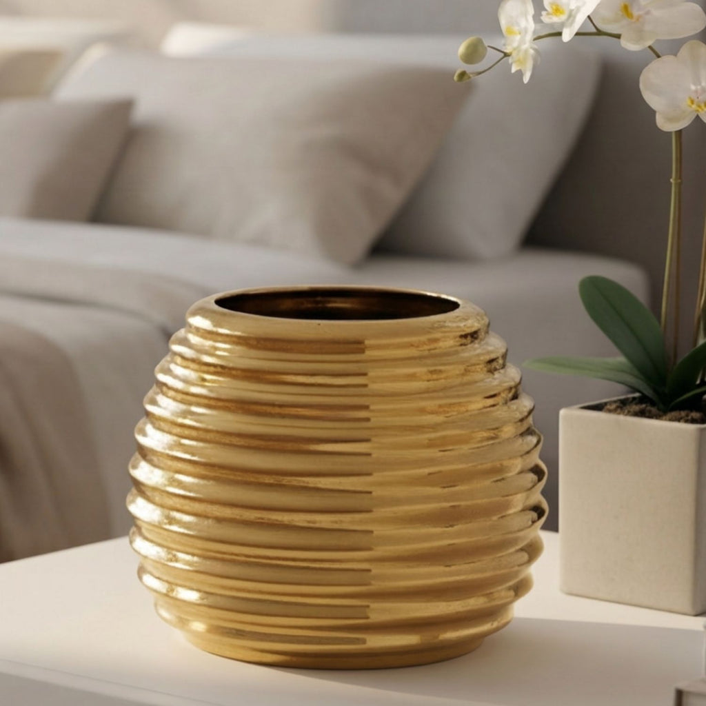 A ribbed, metallic gold ceramic vase sits on a white table in a softly lit living room setting, with a blurred sofa in the background and a potted orchid plant to the right.