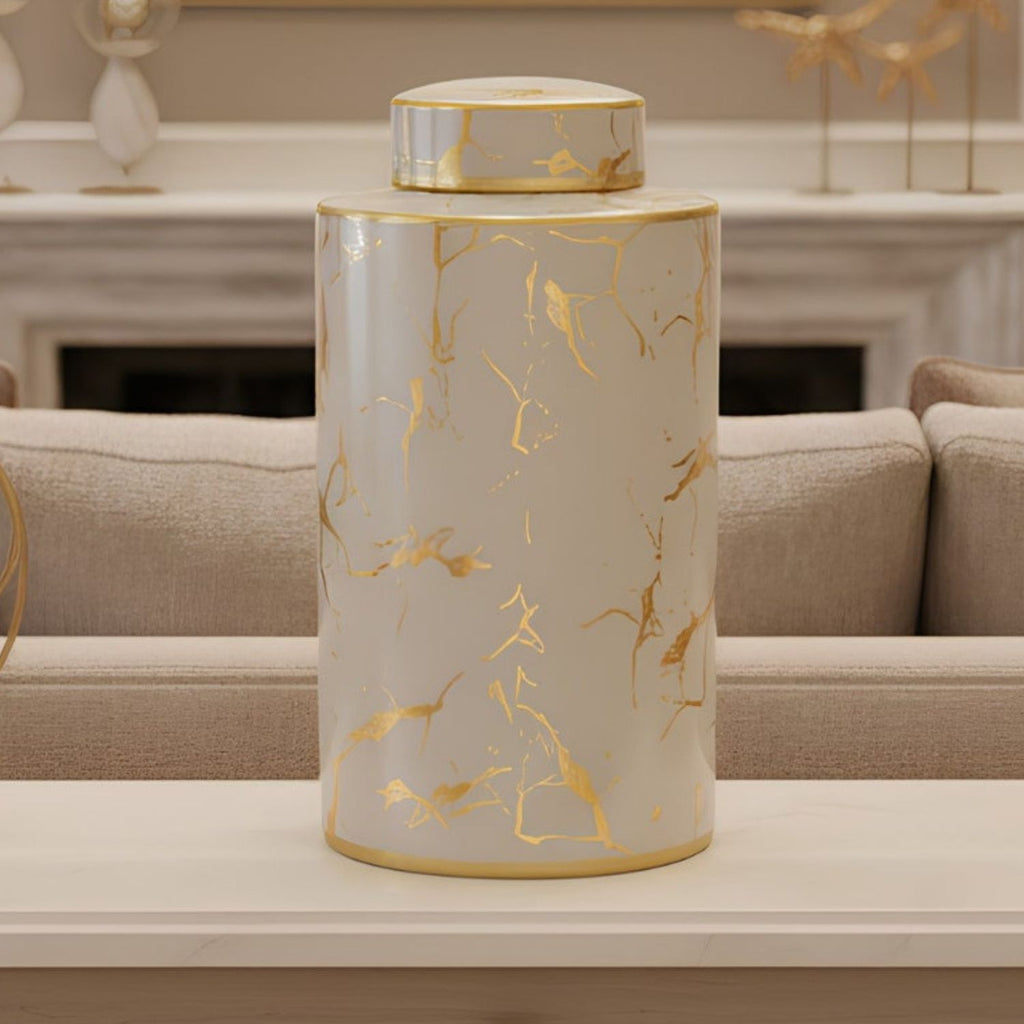 A cylindrical ceramic jar with a matching lid, featuring a white marble pattern with metallic gold veins, displayed on a light-colored table in a modern living room setting.