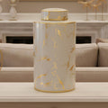 A cylindrical ceramic jar with a matching lid, featuring a white marble pattern with metallic gold veins, displayed on a light-colored table in a modern living room setting.