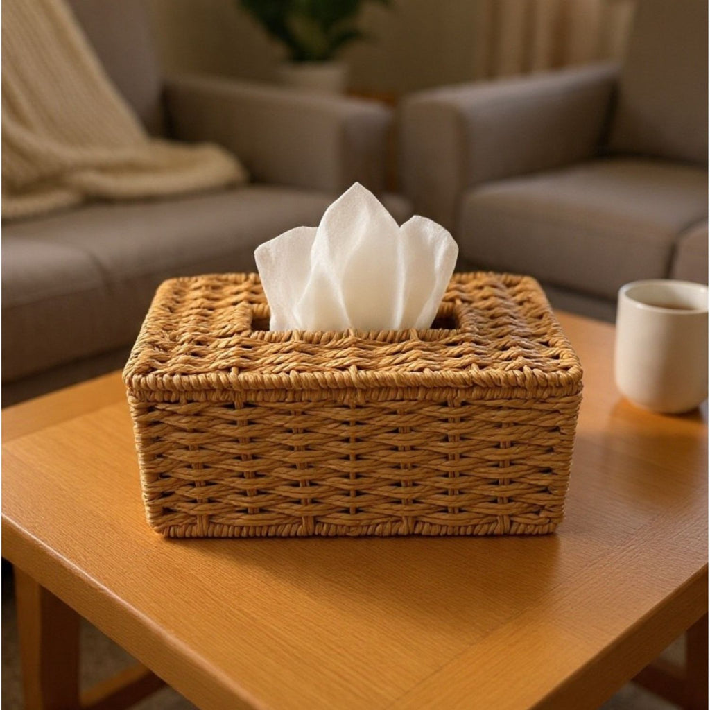 A rectangular woven wicker tissue box cover holding white tissue papers, placed on a wooden table in a softly blurred living room setting with a sofa and mug in the background.