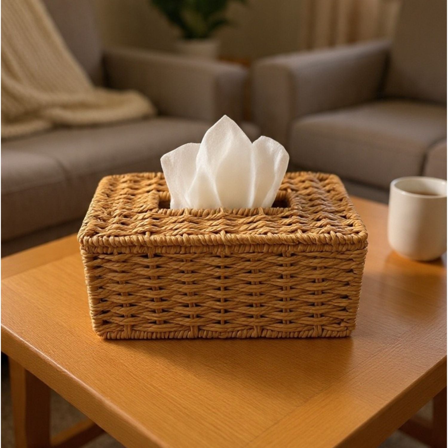 A rectangular woven wicker tissue box cover holding white tissue papers, placed on a wooden table in a softly blurred living room setting with a sofa and mug in the background.