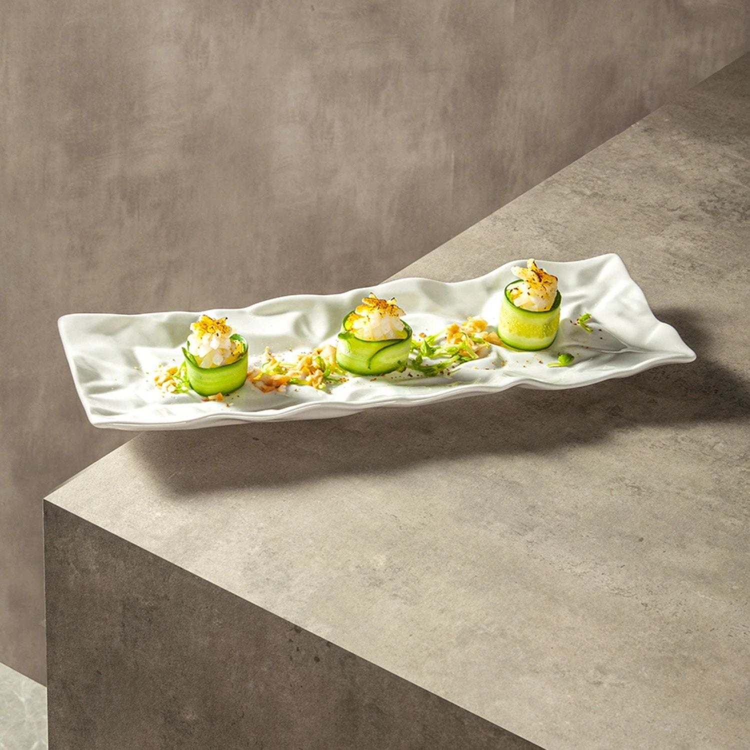 A long, wavy white ceramic serving platter holding three small cucumber appetizers topped with white cream and yellow crumb garnish, set on a gray concrete-style tabletop against a matching gray wall.