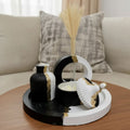 A decorative black and white ceramic tray set with gold accents, styled on a wooden table in front of a beige sofa with a textured pillow. The set includes a small ribbed vase, a crescent-shaped holder with dried grass, a small bowl, and a scalloped shell-shaped decor piece.