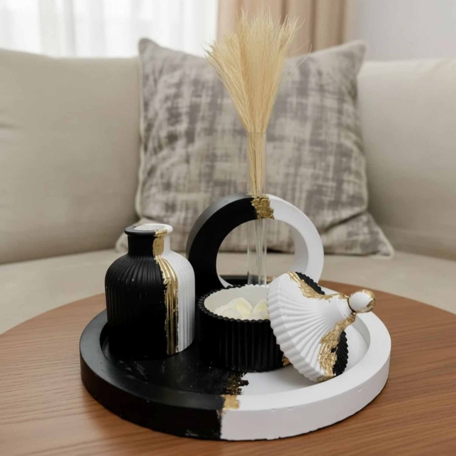 A decorative black and white ceramic tray set with gold accents, styled on a wooden table in front of a beige sofa with a textured pillow. The set includes a small ribbed vase, a crescent-shaped holder with dried grass, a small bowl, and a scalloped shell-shaped decor piece.