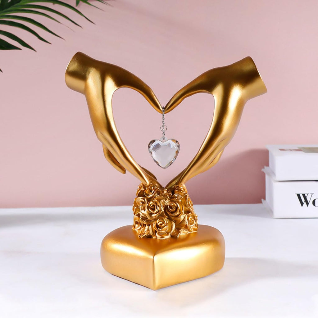 Gold two hands heart sculpture with crystal pendant styled on white surface against pink wall