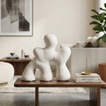 Three abstract white ceramic figurines arranged on a wooden coffee table in a modern, neutral-toned living room setting.