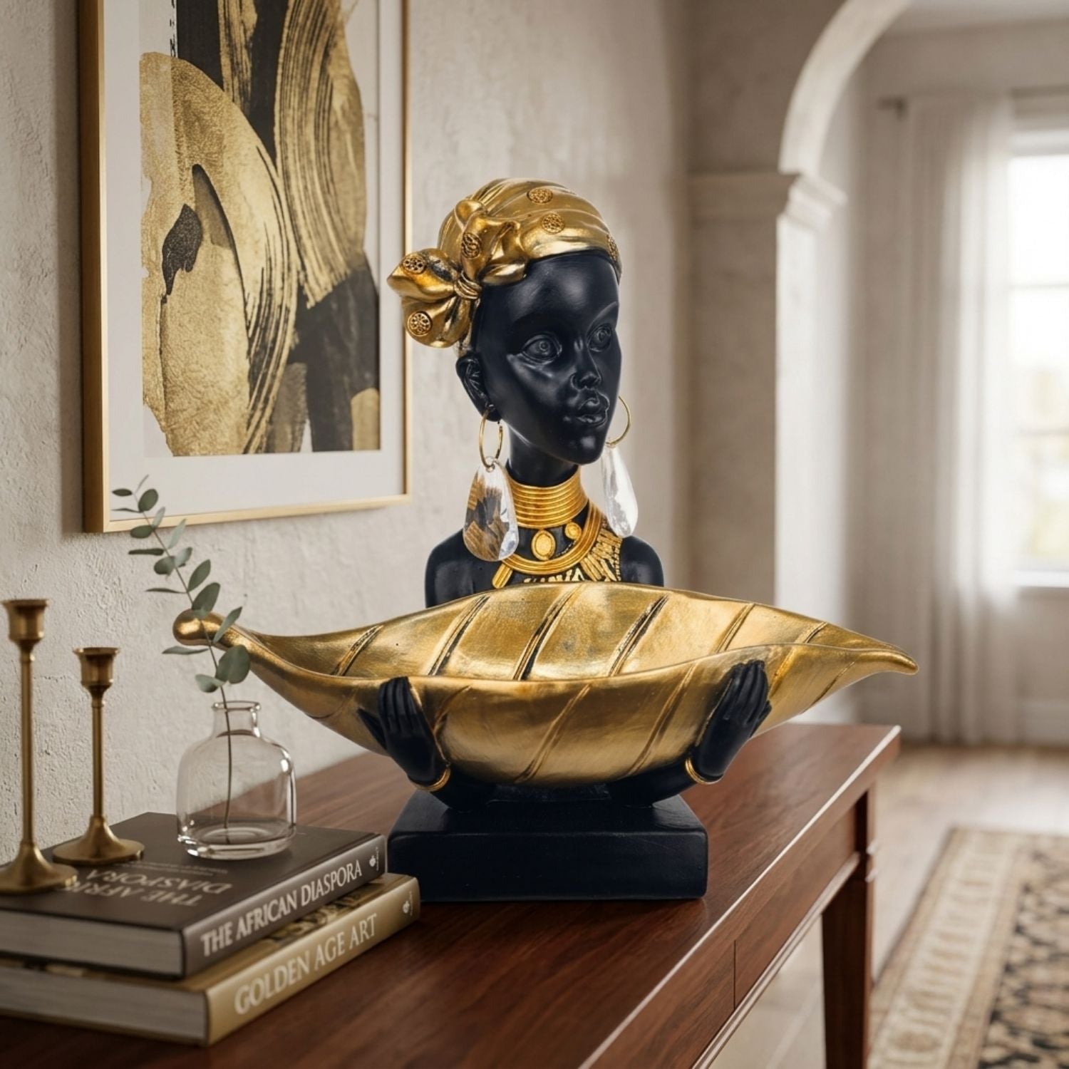 A decorative sculpture of an African woman in a black dress with gold headwrap and jewelry, holding a large gold leaf-shaped bowl, displayed on a wooden console table in a styled interior room.