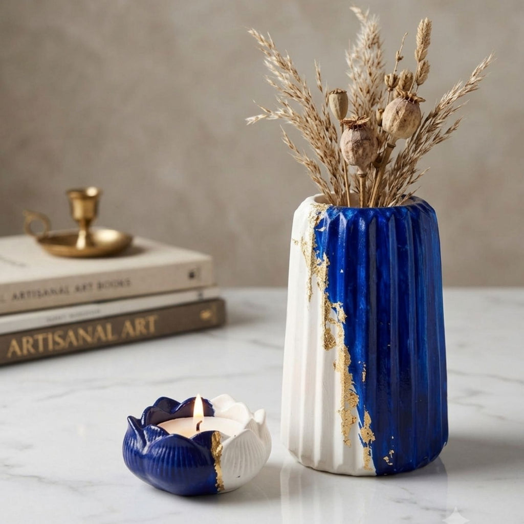 A decorative ceramic vase and matching candle holder in cobalt blue and white with gold leaf accents, styled on a marble surface with dried grasses in the vase and stacked books in the background.