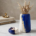 A decorative ceramic vase and matching candle holder in cobalt blue and white with gold leaf accents, styled on a marble surface with dried grasses in the vase and stacked books in the background.