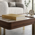A modern coffee table in a styled living room, featuring a dark wood frame, a light marble top, and gold legs. On the table sits a gold book with 'Christian Dior' embossed on the cover, alongside a small glass vase with eucalyptus and a round clock.