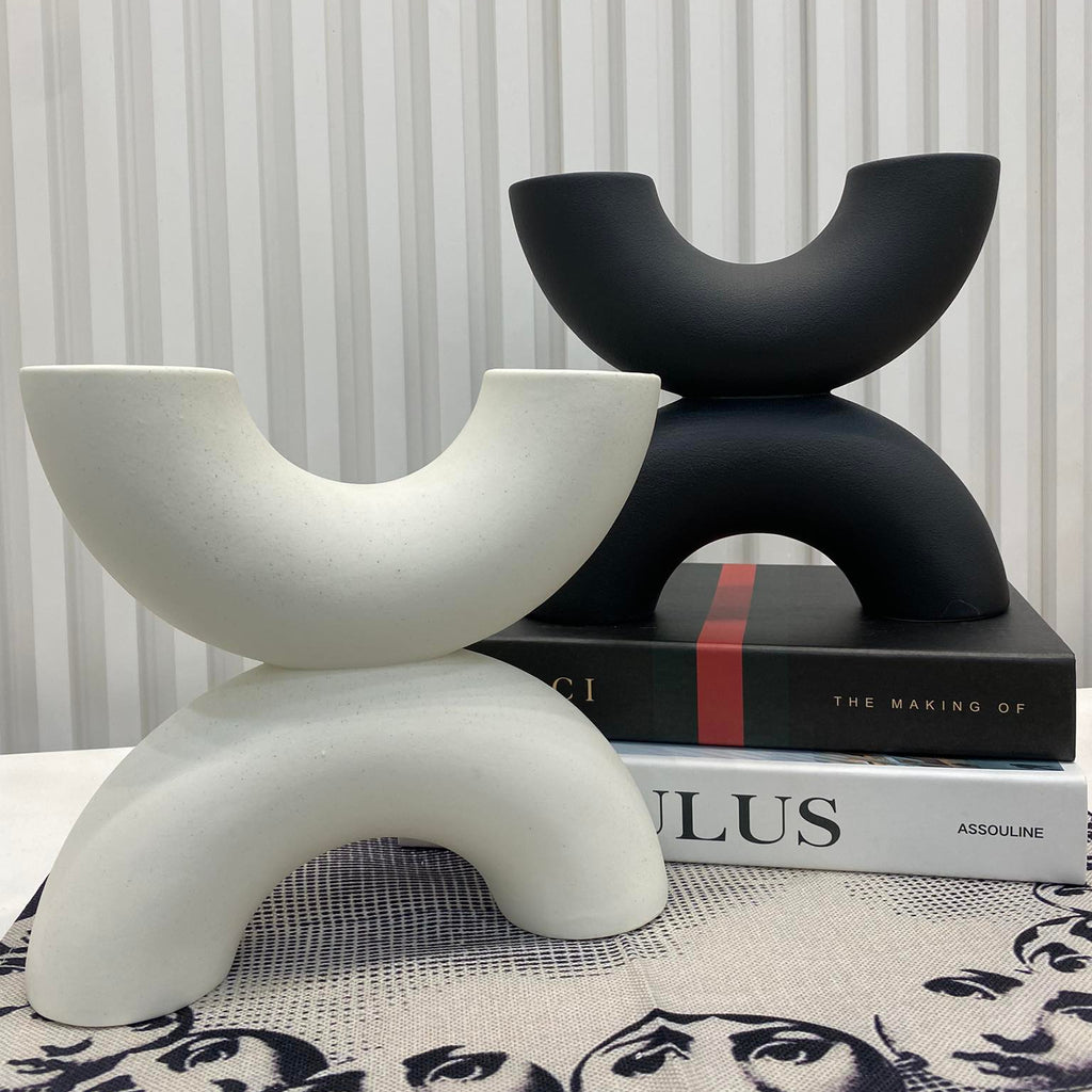 Two modern abstract U-shaped decorative sculptures, one matte white and one matte black, styled on a tabletop with stacked books against a white slatted wall.