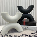 Two modern abstract U-shaped decorative sculptures, one matte white and one matte black, styled on a tabletop with stacked books against a white slatted wall.