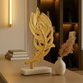 A decorative gold-toned feather sculpture on a rectangular white base, displayed on a tabletop in a styled interior setting with books, a vase, and warm ambient lighting.