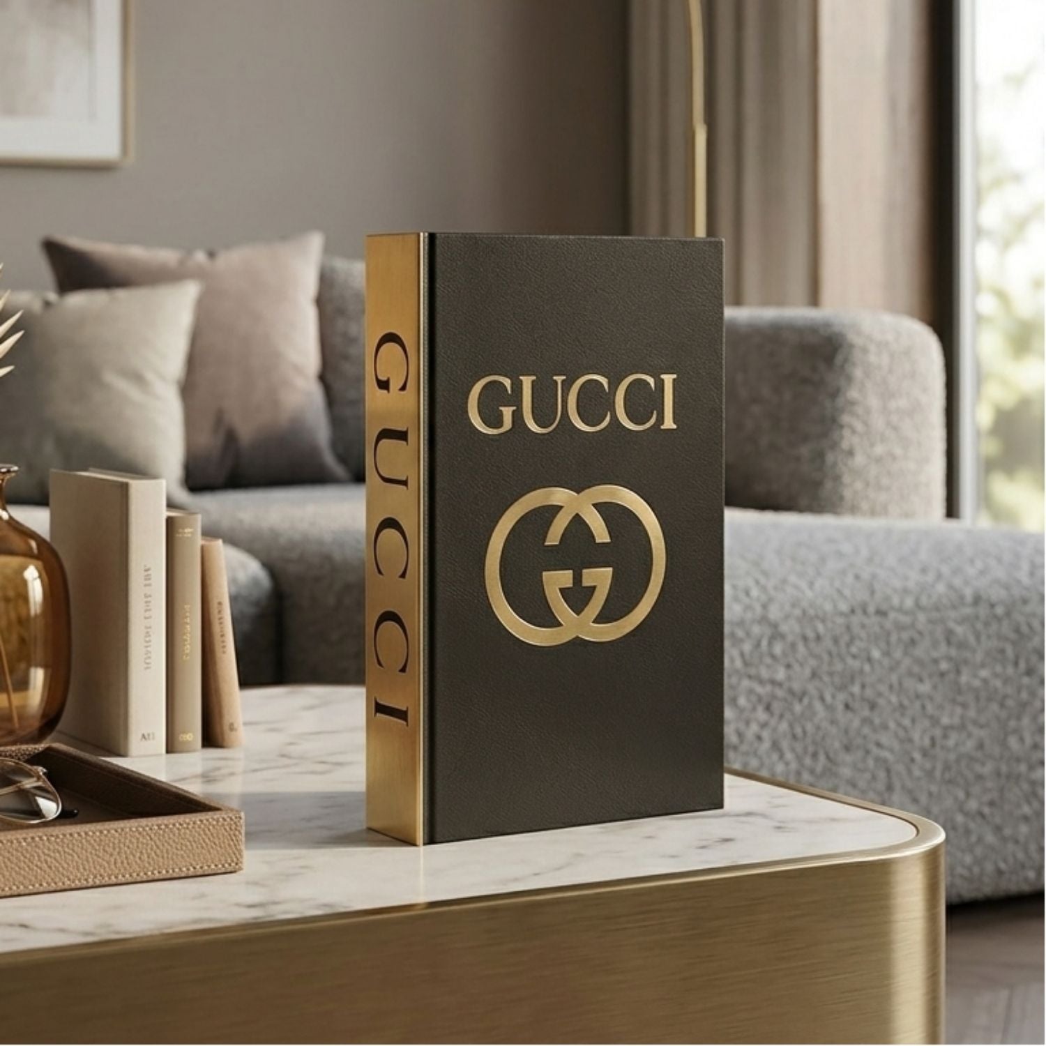 A luxury Gucci hardcover book or decorative book standing upright on a marble-top table in a modern living room setting. The cover is dark charcoal gray with gold lettering reading 'GUCCI' and a large interlocking GG logo, and the spine is gold with vertical 'GUCCI' text.