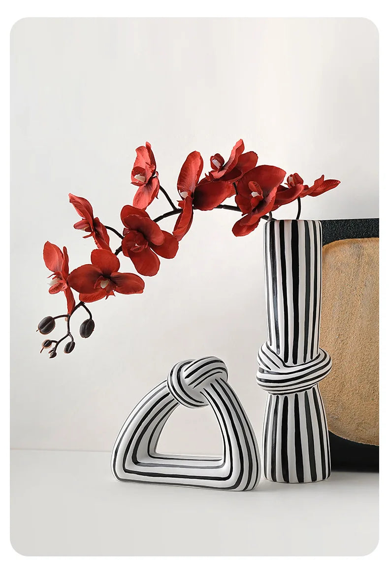 Black and white striped knot vase with red orchids and triangular ornament on white surface with woven hat