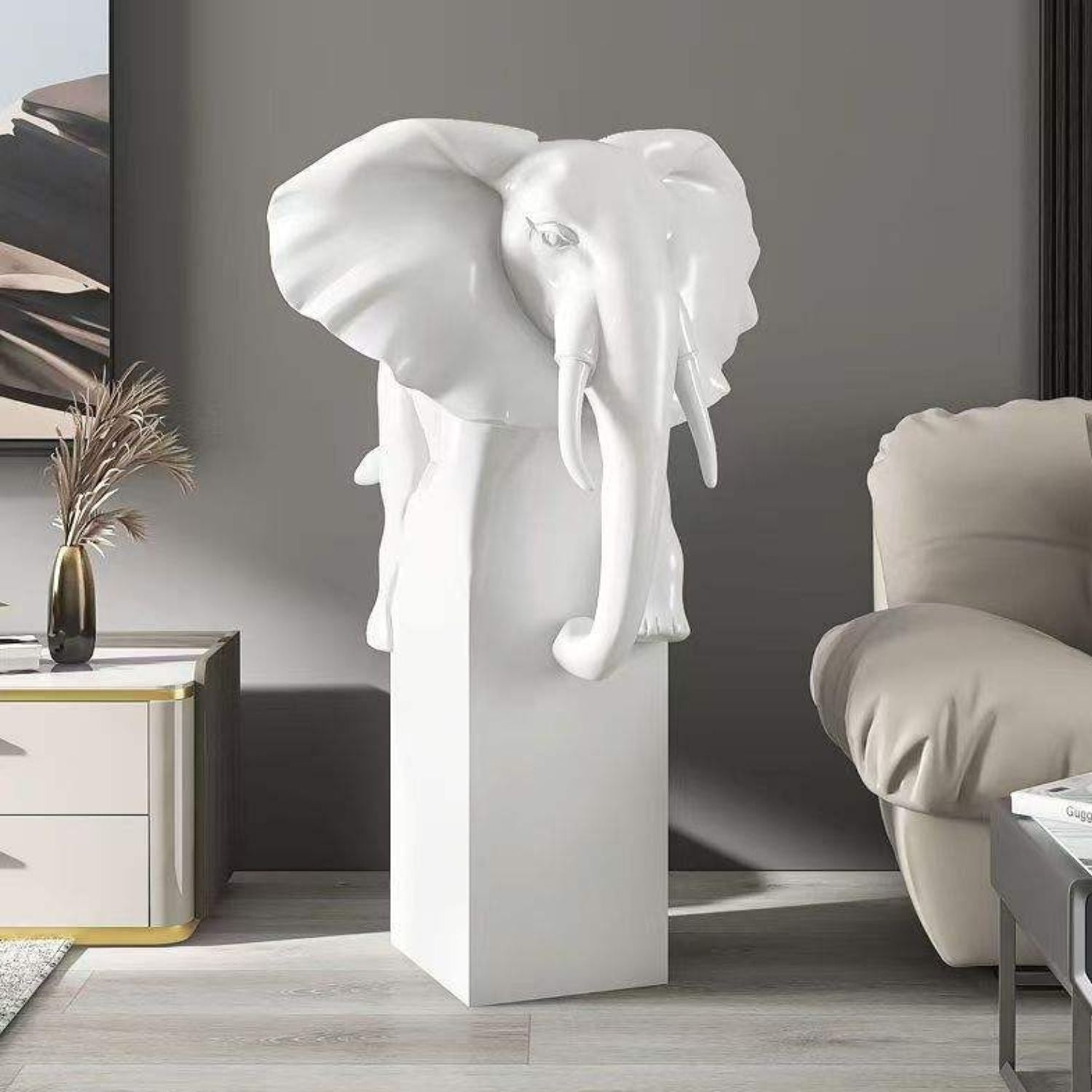 A glossy white elephant head sculpture mounted on a matching white rectangular pedestal, styled in a modern living room setting next to a beige armchair and side table.