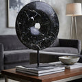 A round black marble donut-shaped table sculpture with white veining, displayed on a black stand on a wooden coffee table in a modern living room setting.
