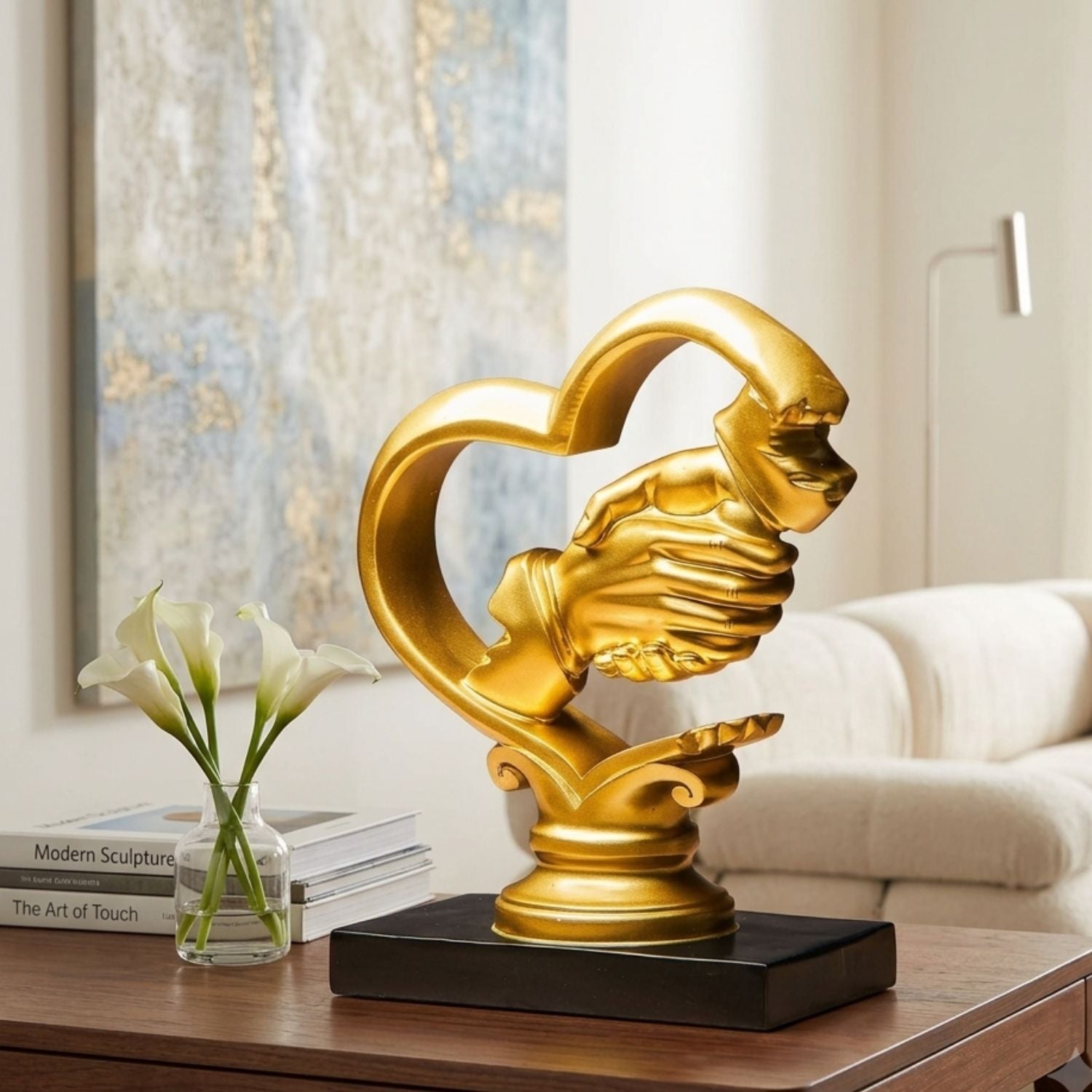 A golden heart-shaped hand sculpture on a black rectangular base, displayed on a wooden table in a modern living room setting next to a vase of white calla lilies and books.
