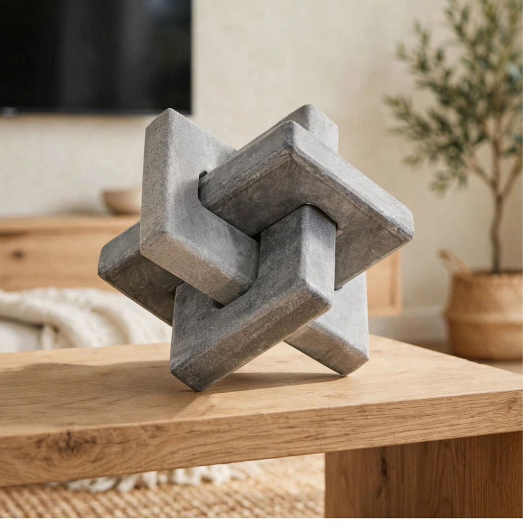 A gray stone-like geometric puzzle sculpture displayed on a light wood table in a softly styled indoor room with blurred furniture and a plant in the background.