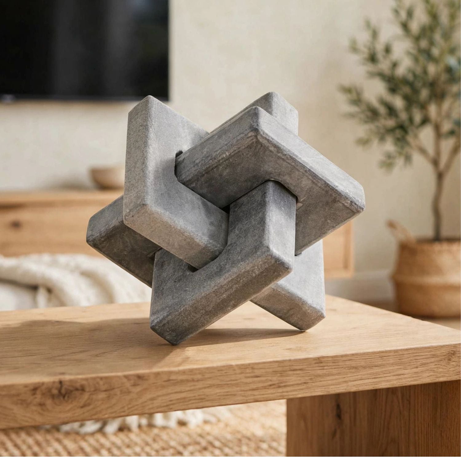 A gray stone-like geometric puzzle sculpture displayed on a light wood table in a softly styled indoor room with blurred furniture and a plant in the background.
