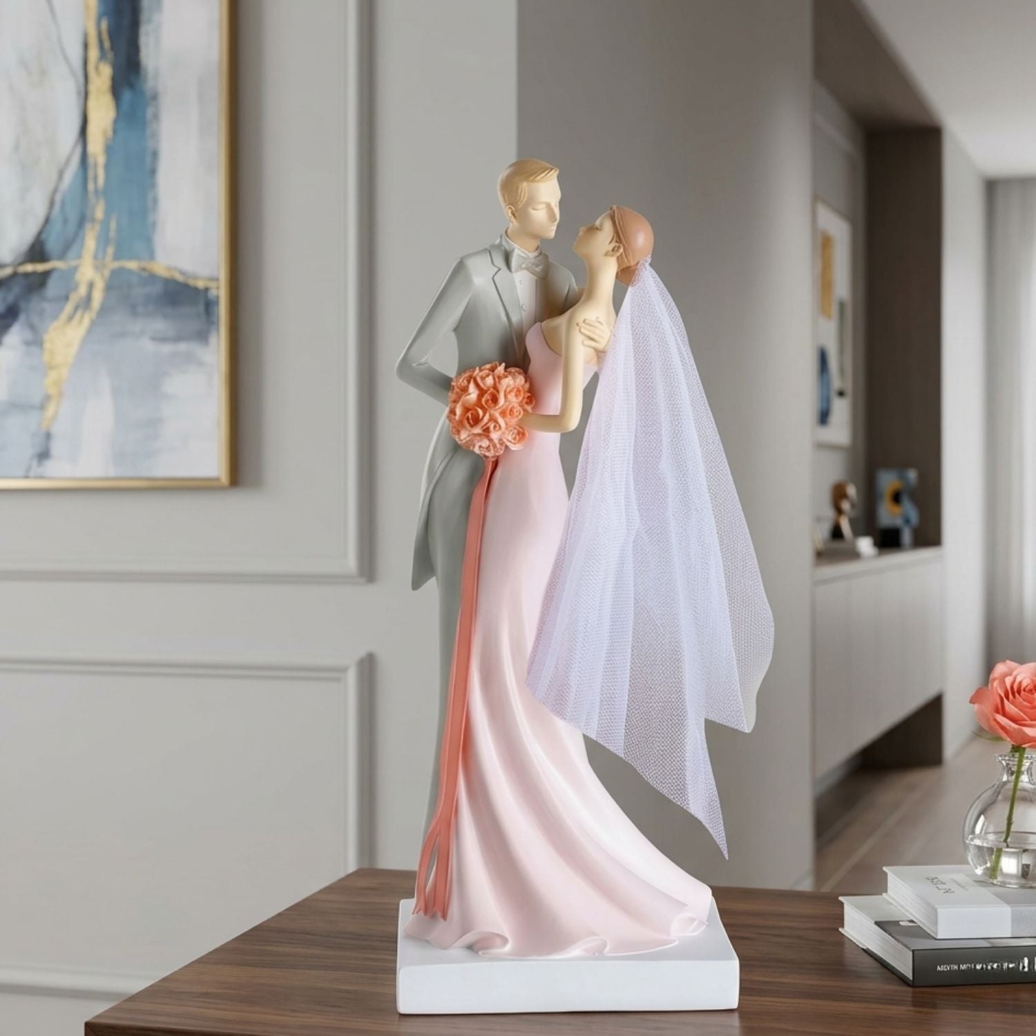 A wedding figurine of a bride and groom embracing, displayed on a wooden table in a modern home interior. The groom wears a gray suit and the bride wears a light pink gown with a white veil and holds a peach-colored bouquet.