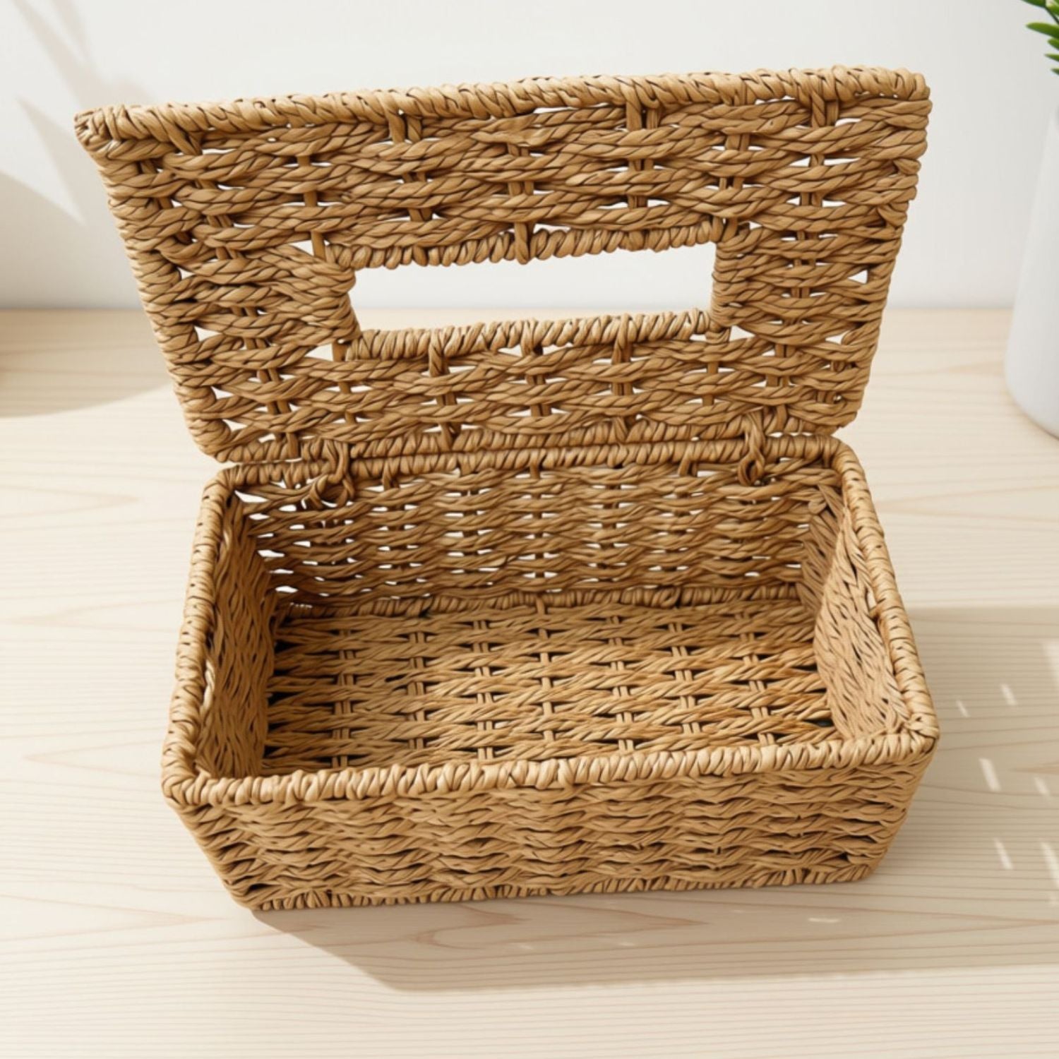 A rectangular woven rattan storage basket with an open hinged lid, shown on a light wood tabletop.
