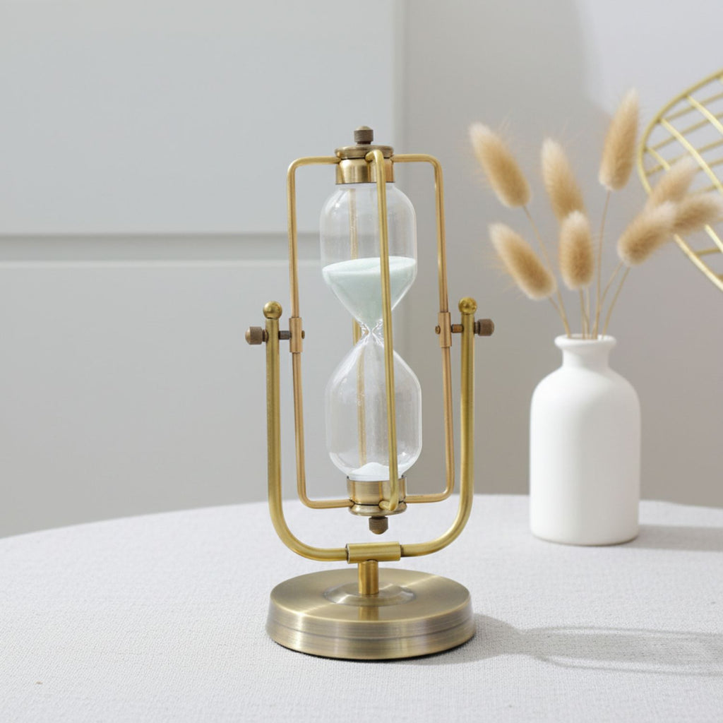 A brass and glass hourglass table lamp with a round base, styled on a light gray table next to a white ceramic vase with dried grass stems.