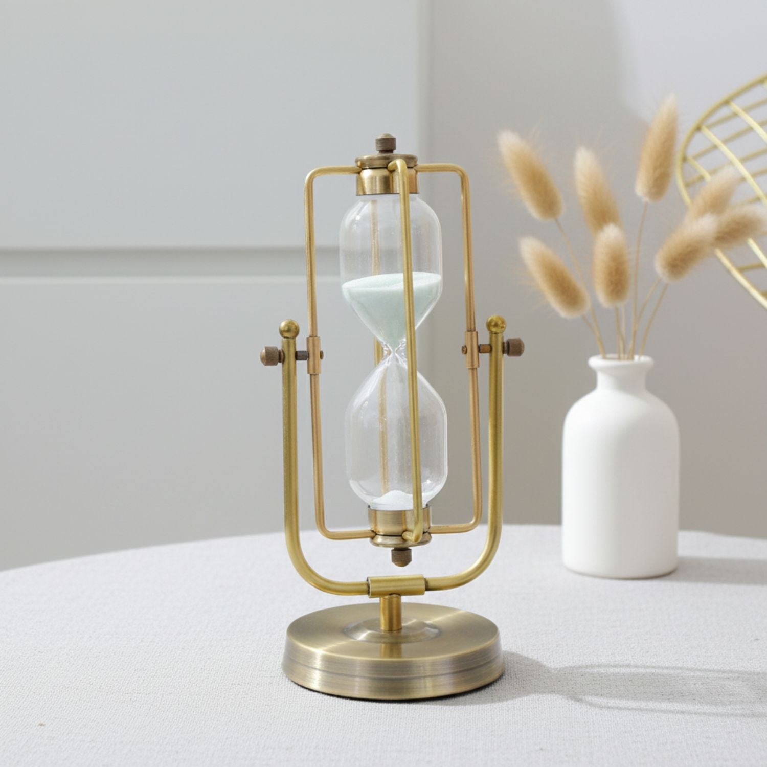 A brass and glass hourglass table lamp with a round base, styled on a light gray table next to a white ceramic vase with dried grass stems.