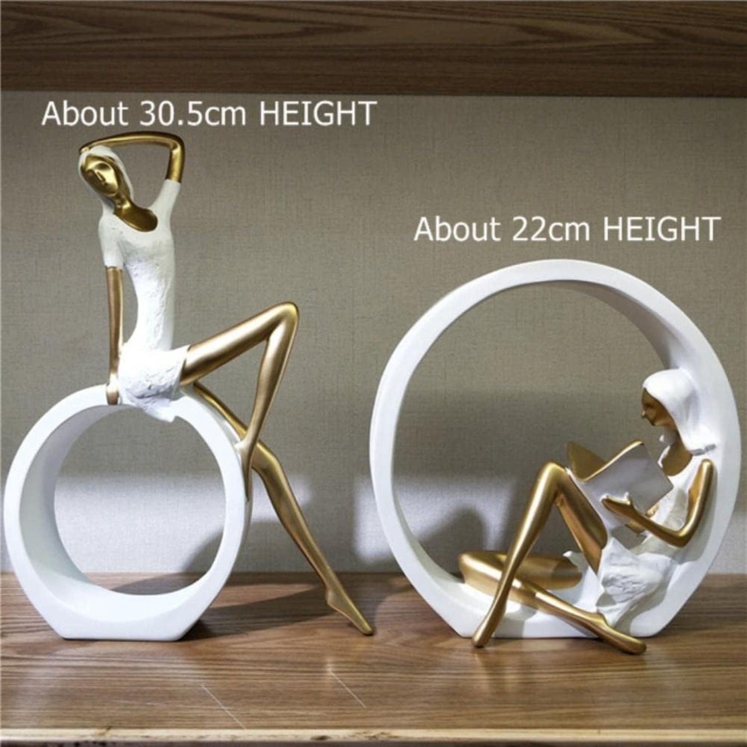 Two white and gold decorative figurines displayed on a wooden shelf against a neutral wall. One figurine sits on a circular base with a standing female form, and the other is a larger circular white ring with a seated female figure in gold.