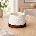 A white ceramic mug with a textured vertical ribbed pattern sits on a round dark brown wooden coaster on a light wood table in a softly blurred home interior.