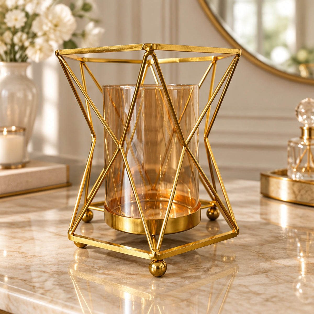A gold geometric metal candle holder with a clear amber-tinted glass cylinder, displayed on a polished marble tabletop in a softly lit interior setting.