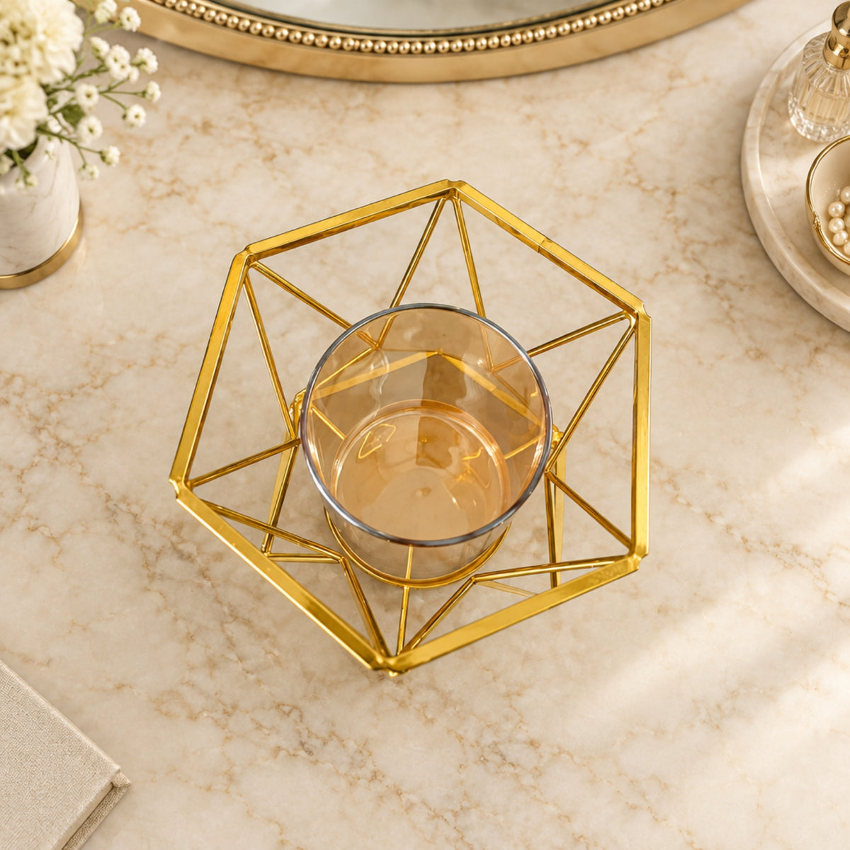A gold geometric metal candle holder with a clear glass candle holder inside, set on a beige marble surface with decorative props.