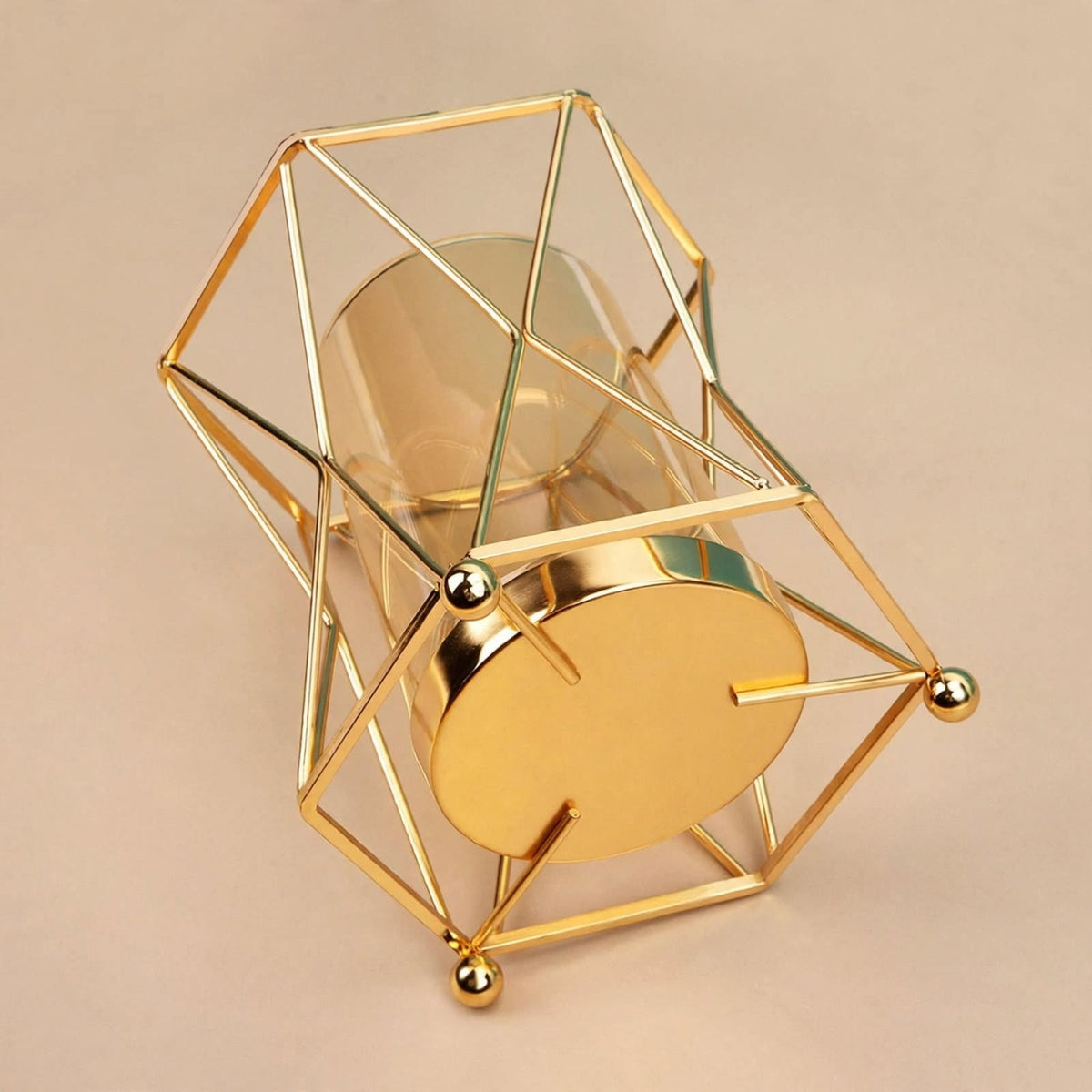 A modern geometric gold metal candle holder with a clear glass cylinder and a round gold base, set against a solid beige background.
