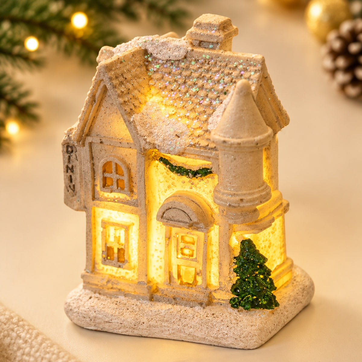 An illuminated Christmas house figurine with a warm yellow glow from within, featuring a textured beige stone-like exterior, glittery roof, and a small green tree, shown on a softly lit holiday background.