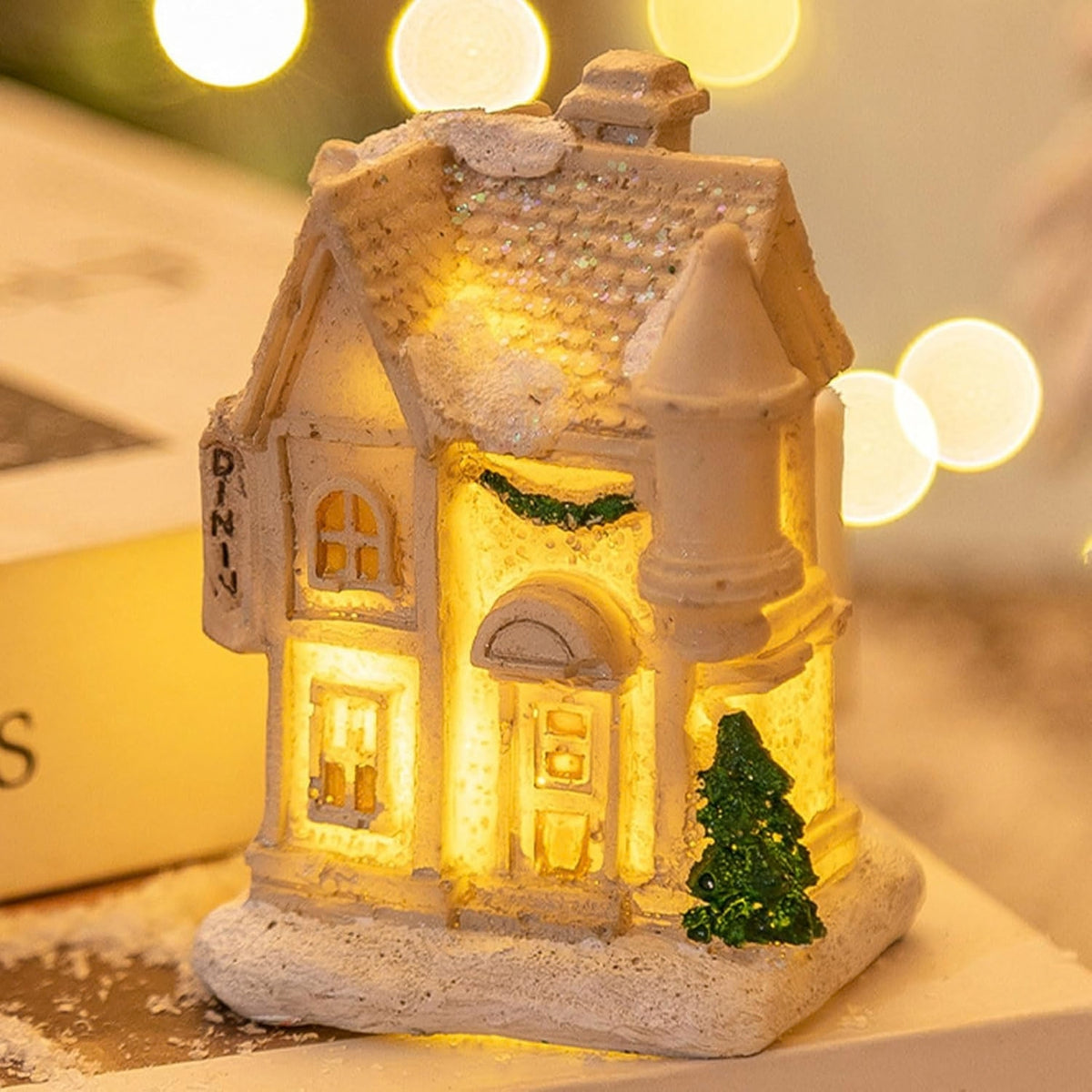 A small illuminated Christmas house figurine with warm yellow lights glowing from within, a snow-dusted roof, a green tree accent, and a vertical 'DININ' sign on the side, photographed on a tabletop with blurred warm lights in the background.