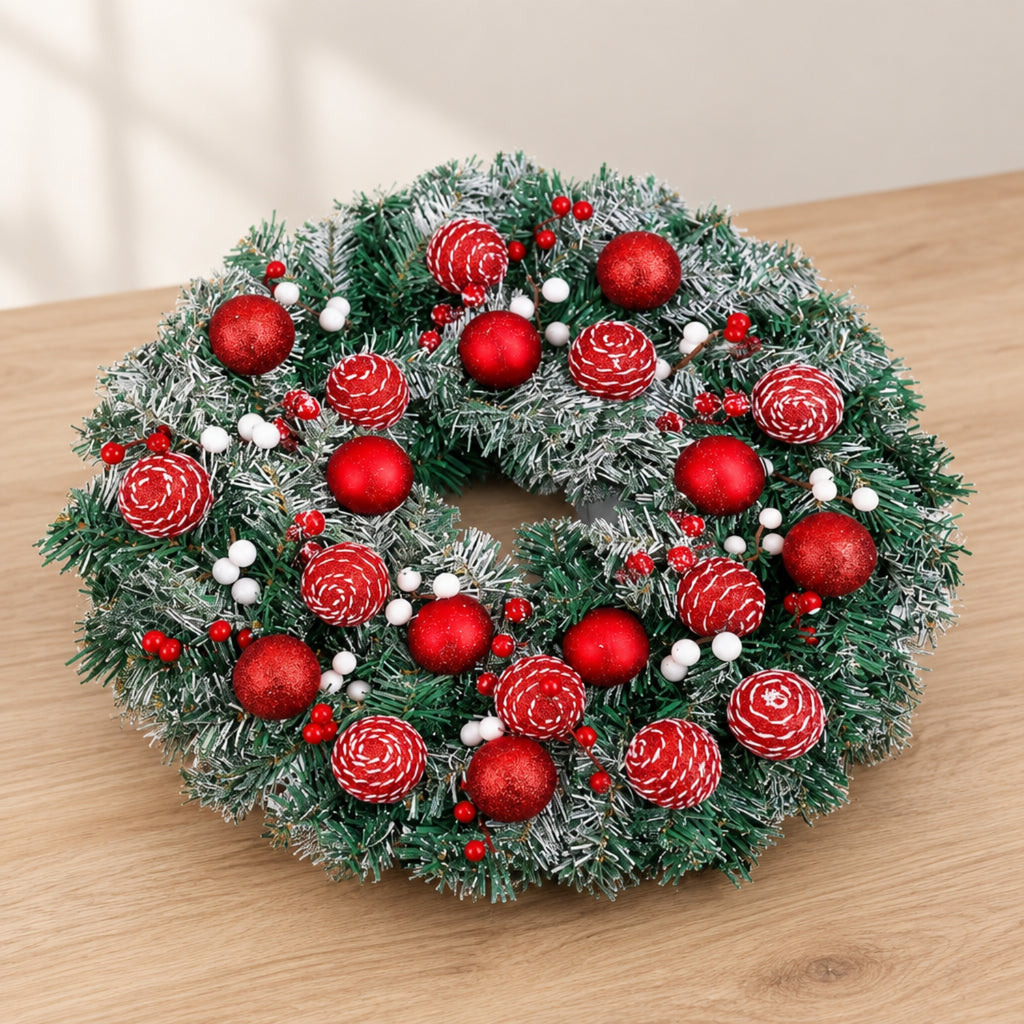 A festive artificial Christmas wreath with green frosted pine needles, decorated with red ornaments, white berries, and pinecones, displayed on a light wood tabletop.