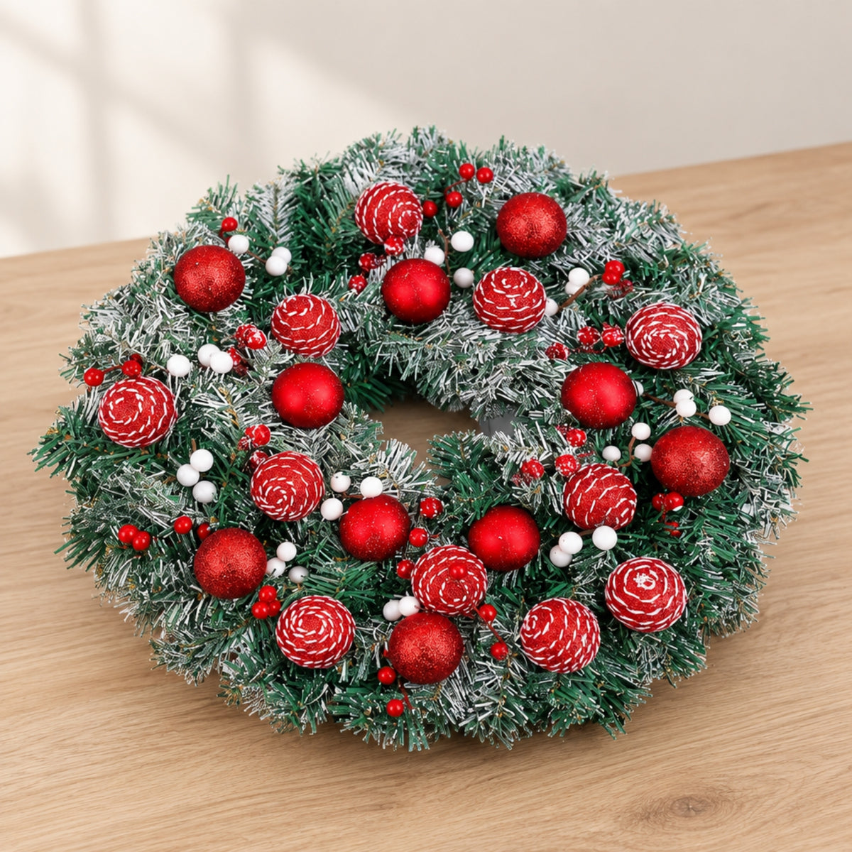 A festive artificial Christmas wreath with green frosted pine needles, decorated with red ornaments, white berries, and pinecones, displayed on a light wood tabletop.