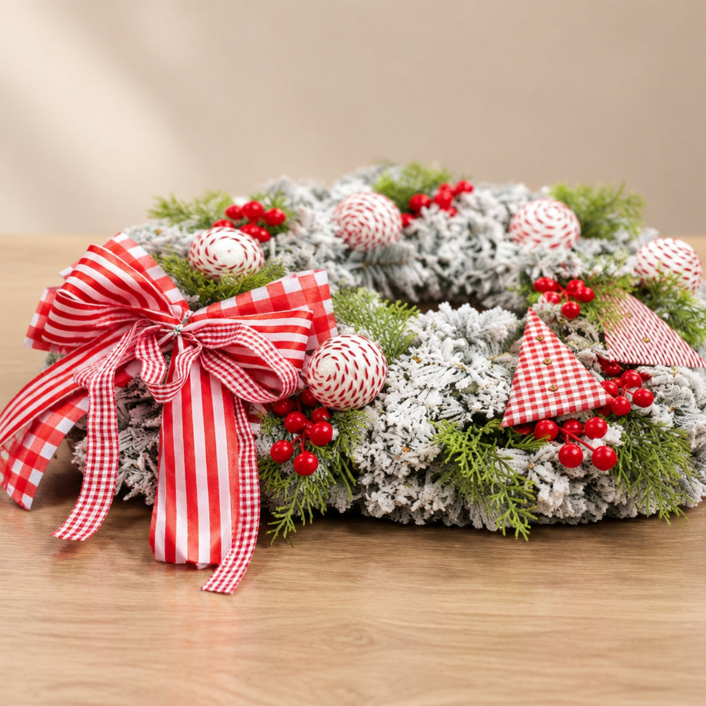 A frosted Christmas wreath with green pine branches, red berry clusters, white gingham ribbon ornaments, and a large red-and-white striped bow, displayed on a wooden surface against a warm beige background.