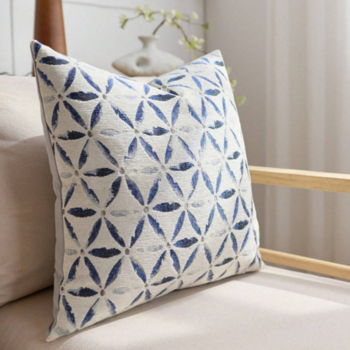 A square throw pillow with a blue and white geometric leaf pattern, resting on a beige sofa in a softly lit room.