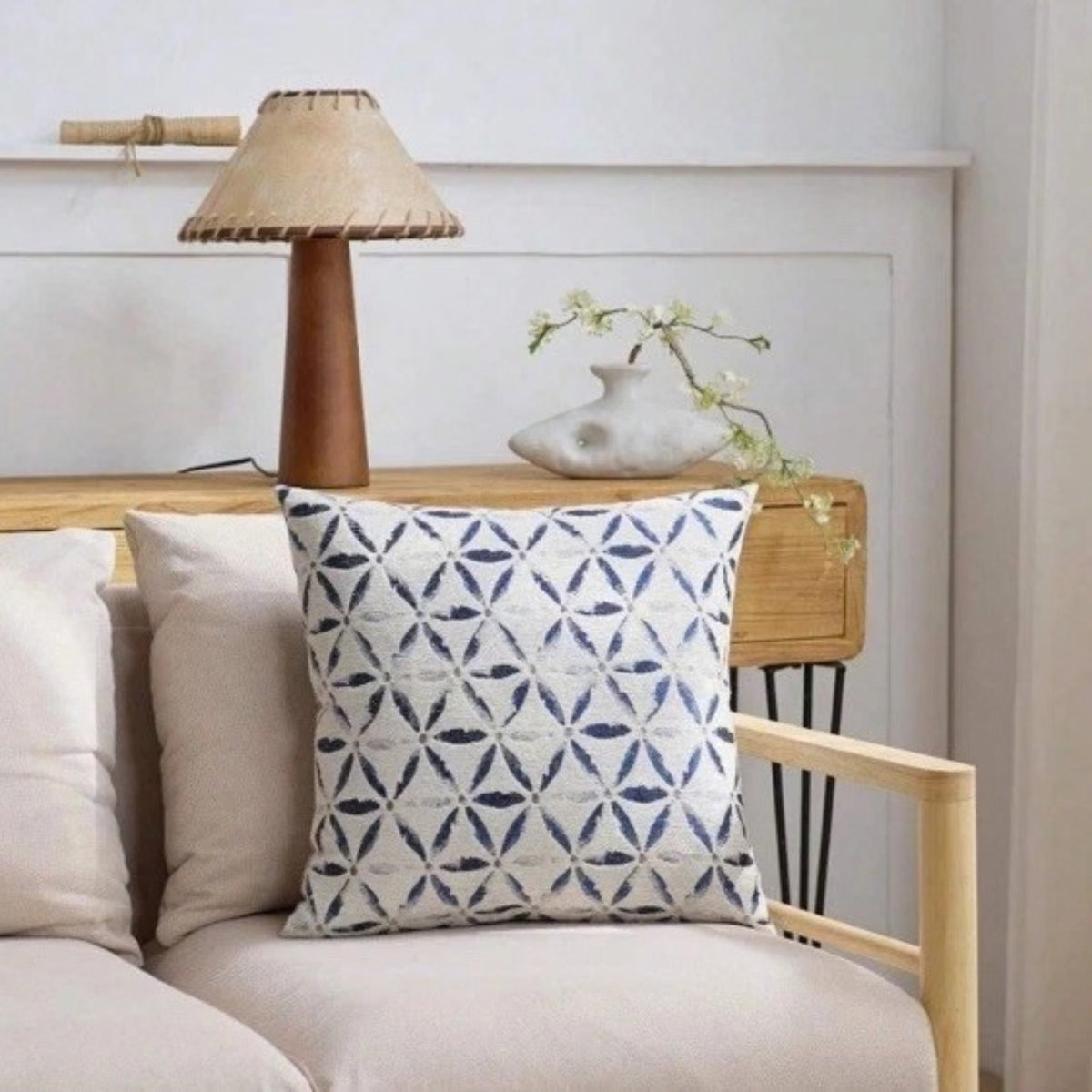 A square throw pillow with a blue and white geometric pattern, styled on a light beige sofa next to a wooden side table with a lamp and a small vase.