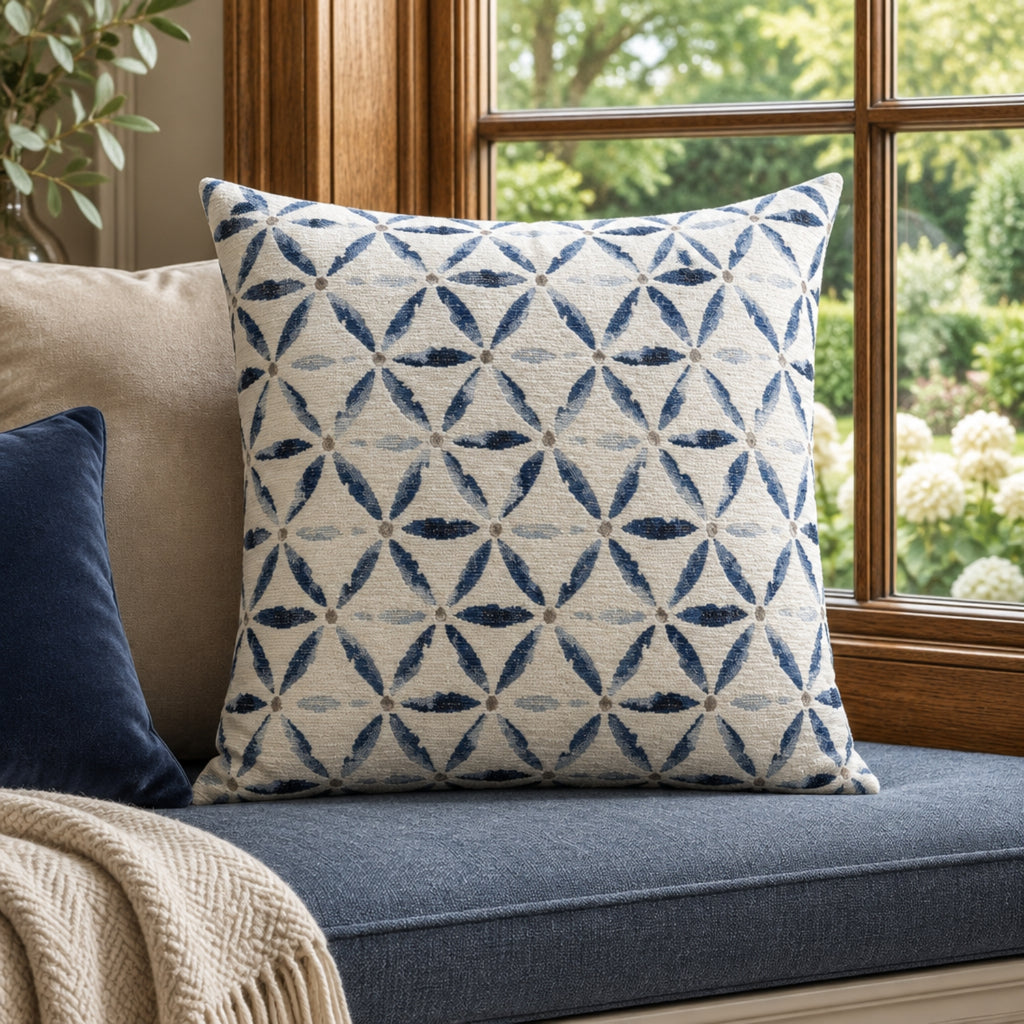 A square throw pillow with a blue geometric trellis pattern on a cream background, styled on a blue window seat cushion in a sunlit room.