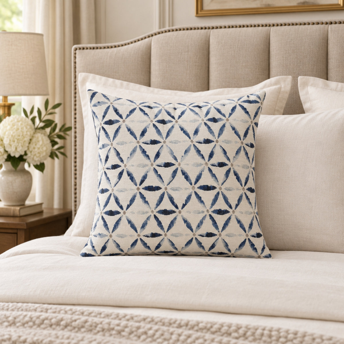 A square decorative throw pillow with a blue and white geometric pattern, styled on a cream-colored bed with a tufted headboard.