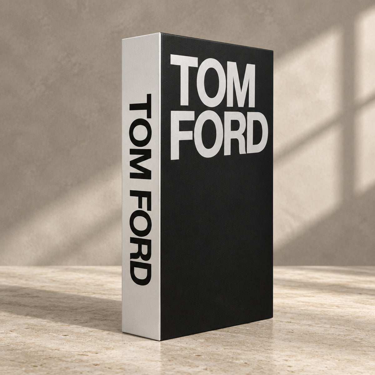 A tall rectangular book or box standing upright on a stone-like surface, with a black front cover and white side panel. Large white text on the front reads TOM FORD, and the spine also reads TOM FORD.