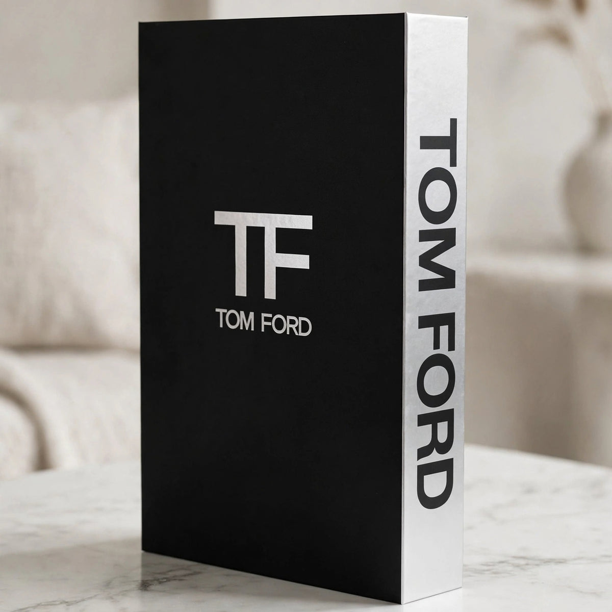 A black and white Tom Ford hardcover book standing upright on a marble surface with a softly blurred indoor background. The front cover is black with a large white 'TF' logo and 'TOM FORD' text, while the spine is white with 'TOM FORD' printed vertically in black.