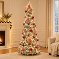 A tall flocked Christmas tree decorated with red ornaments, cream ribbons, and warm white string lights, standing in a cozy living room with a fireplace, curtains, and a sofa.