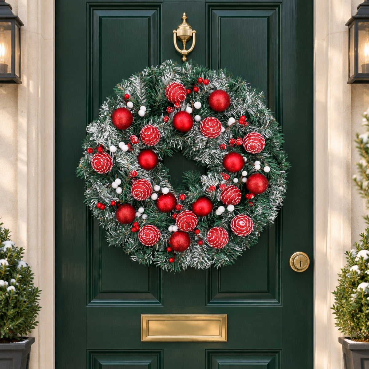 A festive Christmas wreath with red ornaments, pinecones, and white berries hanging on a dark green front door, flanked by potted plants and wall lanterns.