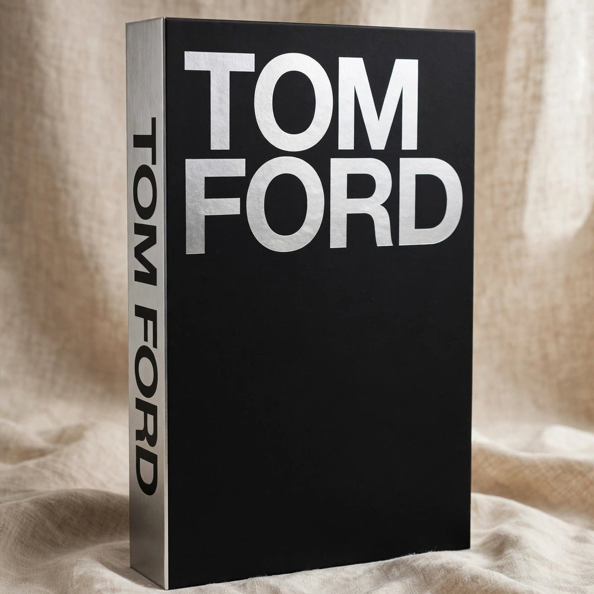 A hardcover book with a black cover and silver spine, standing upright on draped beige fabric. The front cover features the name 'TOM FORD' in large silver letters, and the spine also reads 'TOM FORD'.
