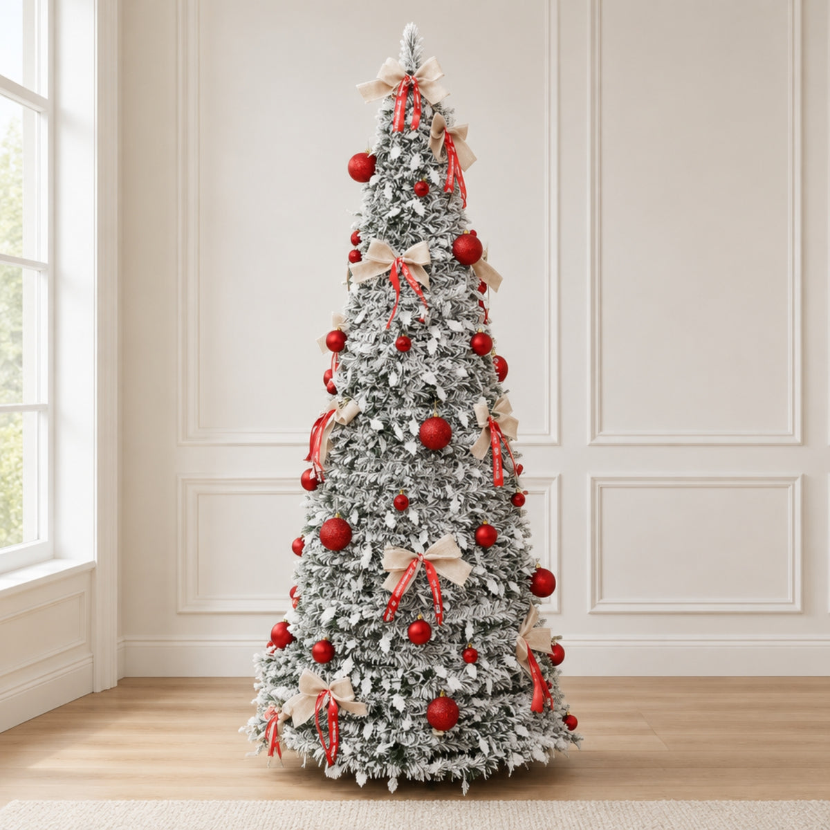 A tall, flocked Christmas tree decorated with red ornaments and beige ribbon bows, standing on a light wood floor against a cream paneled wall.